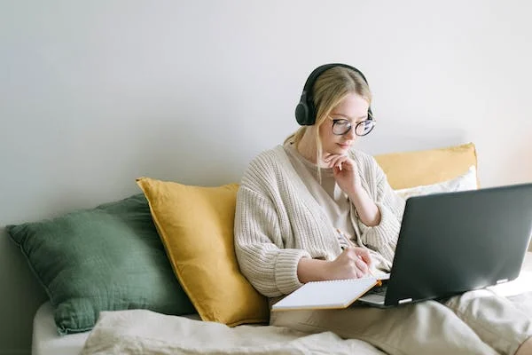 female sitting on bed on laptop with notebook with headphones on